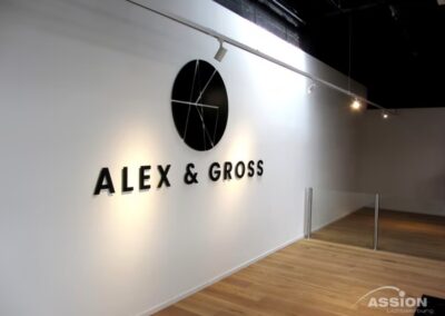 Eine weiße Wand mit einem schwarzen kreisförmigen Logo und dem Text "ALEX & GROSS" in schwarzen Buchstaben; Holzboden und Scheinwerfer über dem Kopf.