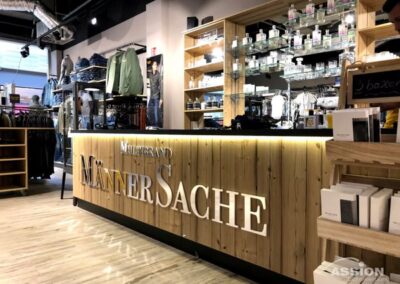 Bekleidungsgeschäft mit einer hölzernen Theke mit der Aufschrift "Männersache", Regalen mit gefalteter Kleidung, hängenden Jacken und Glasregalen mit Pflegeprodukten.