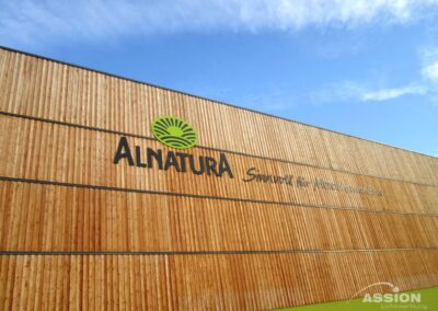Das Äußere eines Alnatura Gebäudes zeigt eine Holzfassade mit dem Alnatura Logo und dem Slogan unter einem blauen Himmel.