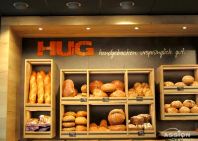 Eine Bäckereiauslage mit mehreren Regalen mit verschiedenen Brotsorten und Brötchen. An der Wand steht das Wort "HUG" und deutscher Text über den Regalen.