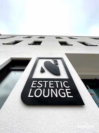 Ein Gebäude mit einem Schild mit der Aufschrift "ESTETIC LOUNGE" und einem minimalistischen Gesichtslogo an einer weißen Wand.