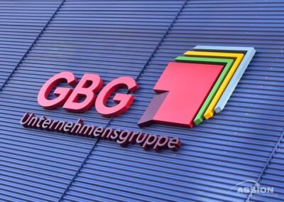 Logo und Name der GBG Unternehmensgruppe auf einer blauen Wellblechwand, mit einem mehrschichtigen, bunten geometrischen Symbol neben dem Text.