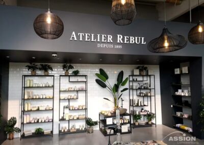 Regale mit Schönheitsprodukten säumen die Wand unter dem Schild "Atelier Rebul Depuis 1895" in einem modernen, gut beleuchteten Ladeninneren mit Hängeleuchten und Topfpflanzen.