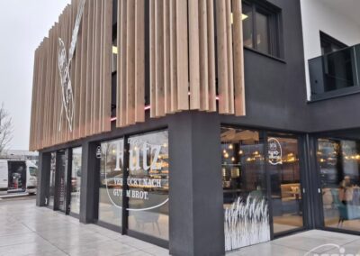 Modernes zweigeschossiges Gebäude mit großen Fenstern mit eleganten Folienbeklebungen und vertikalen Holzlamellen an der Fassade; Innenbeleuchtung und Menschen durch das Glas sichtbar.