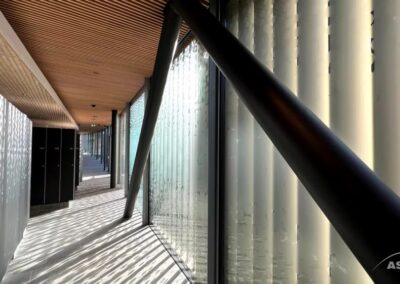 Ein moderner Flur mit Glaswänden, vertikalen Jalousien, schwarzen Strukturbalken, Sonnenlichtmustern auf dem Boden und einer Holzlattendecke.