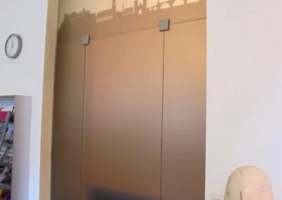 Aufzugstüren aus Milchglas mit der Skyline einer Stadt im oberen Bereich, daneben eine Uhr, Broschüren und ein skulpturales Objekt in einem modernen Interieur.