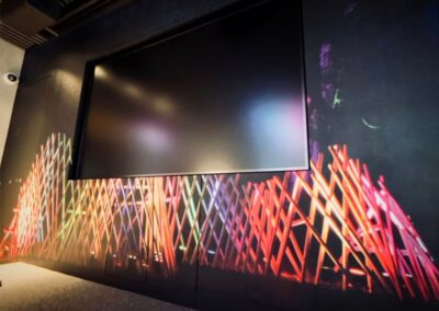 An einer dunklen Wand ist ein großer leerer Bildschirm angebracht, der mit bunten, sich kreuzenden Lichtmustern verziert ist; in der Ecke ist das Logo der ASSION Lichtwerbung zu sehen.