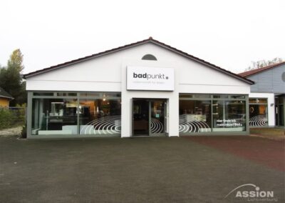 Ein modernes Ausstellungsgebäude mit großen Glasfenstern, einem "badpunkt"-Schriftzug über dem Eingang und weißen grafischen Mustern auf den Fenstern.