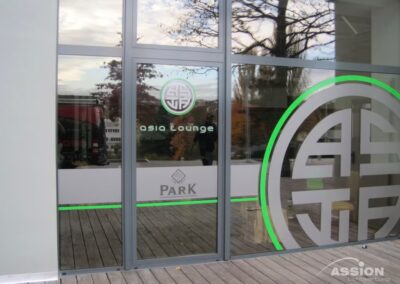 Gläserne Eingangstür zur Asia Lounge mit Logo und "PARK"-Schild, daneben eine Holzterrasse, in deren Glas sich Bäume und Gebäude spiegeln.