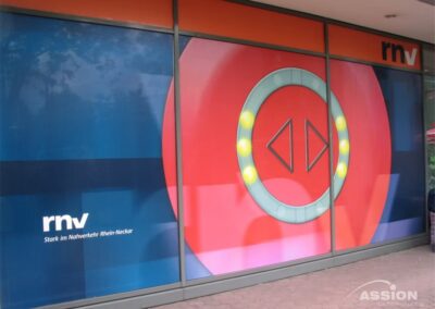 Ein Schaufenster zeigt das Logo und den Slogan der rnv in Form einer großen Grafik mit kreisförmigem Design und den Farben Rot, Blau und Orange.