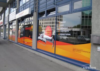 In den Schaufenstern ist eine große Werbung für eine Therapieklinik zu sehen, die Dienstleistungen wie Massage, Akupunktur, Chiropraktik und Infrarotwärme auflistet und eine Person zeigt, die auf einer Therapieliege liegt.
