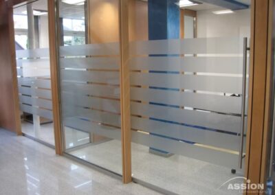 Bürotrennwände aus Glas mit horizontalen, mattierten Streifen, Holzrahmen und einer blauen Säule in einer modernen Büroeinrichtung.